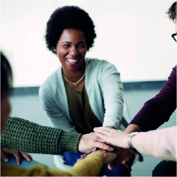 A group of people sit in a circle, placing their hands on top of each other's in the center. One person is smiling.