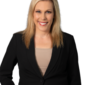 Blonde woman in a black blazer over a beige top, smiling against a dark blue patterned background.