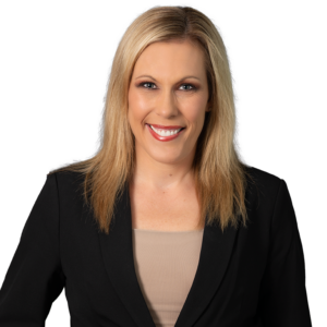Blonde woman in a black blazer over a beige top, smiling against a dark blue patterned background.