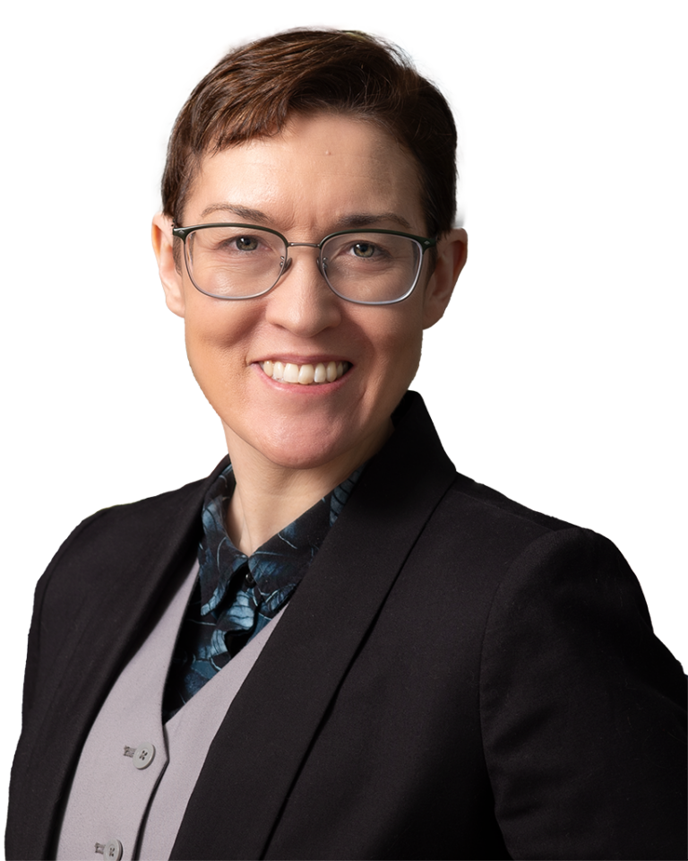 A person with short hair and glasses wearing a suit jacket and vest smiles at the camera, standing in front of a colorful, abstract background.