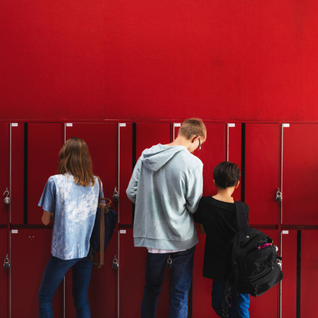 Five students stand in front of red lockers, some opening them and others facing away, against a solid red wall background—a scene reminiscent of environments where compliance with the Public Records Act is essential.