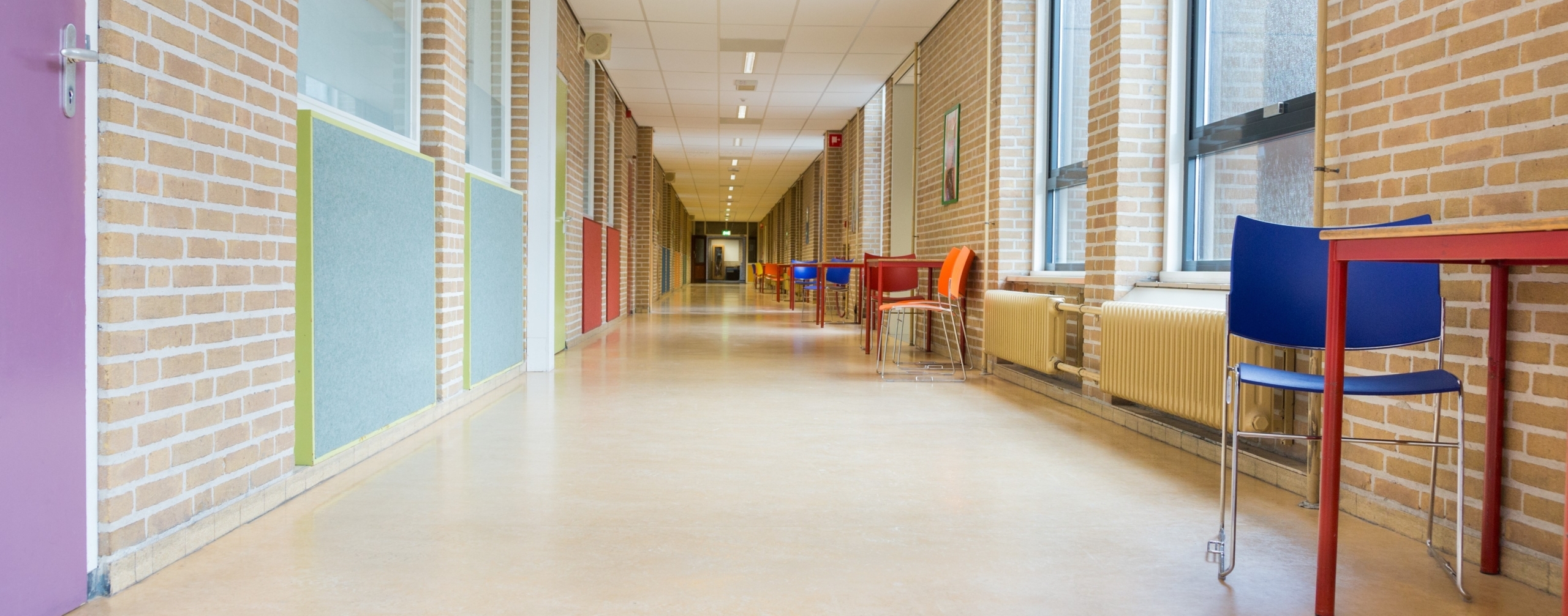 A long, empty hallway in a school or office building awaiting charter renewal, with brick walls, large windows, and colorful chairs and tables lining one side.