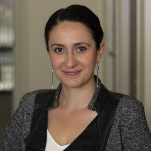 Woman with dark hair tied back, wearing a grey blazer with black details and long earrings, stands indoors and smiles at the camera, embodying professionalism in wage and hour compliance.