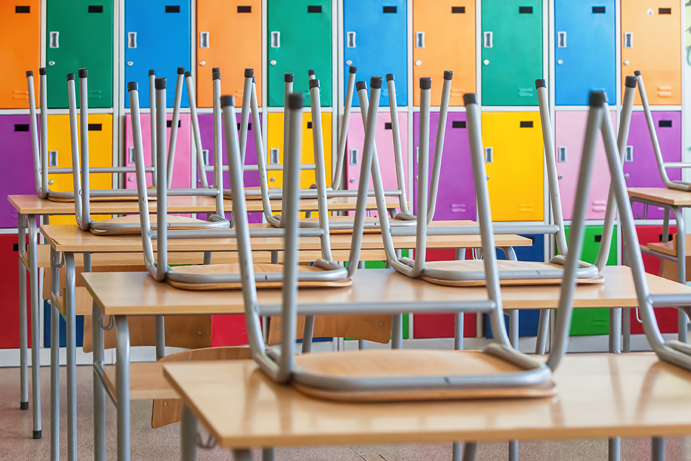 Empty classroom with colorful lockers in the background and chairs stacked upside down on wooden desks, reflecting efforts to reduce risk and manage crisis in educational spaces.