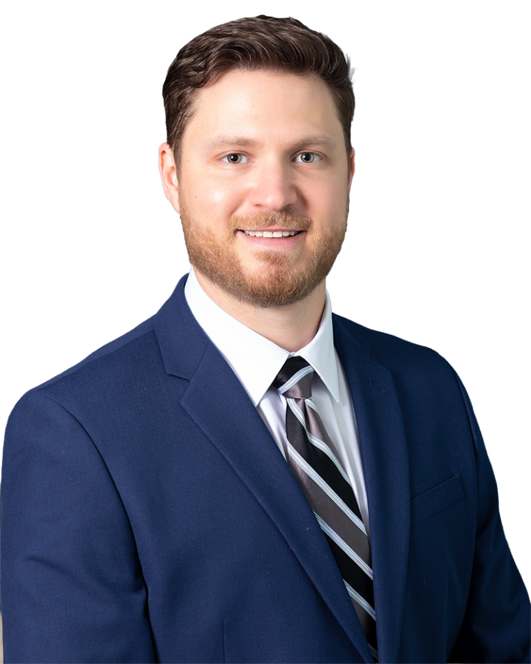 Man with short brown hair and beard wearing a navy blue suit, white dress shirt, and striped tie, standing against a plain white background, facing the camera and smiling.
