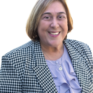 Smiling person with short light brown hair wears a black and white houndstooth blazer, lavender blouse, and a Star of David necklace, standing against a plain white background.