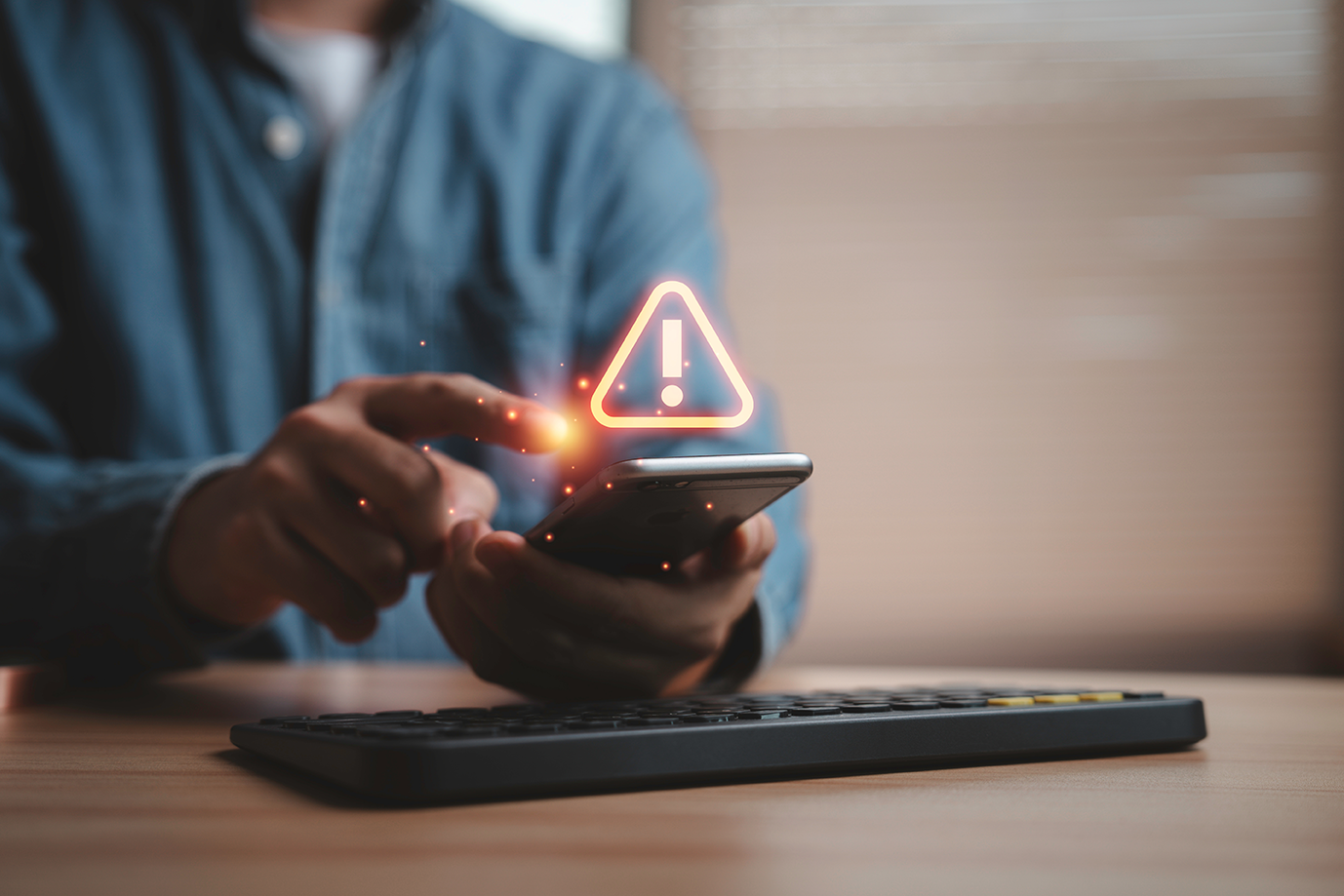 A person uses a smartphone, with a glowing fraud alert symbol appearing above the device—possibly signaling an invoice payment scam or law firm impersonation attempt; a keyboard is on the table in front of them.
