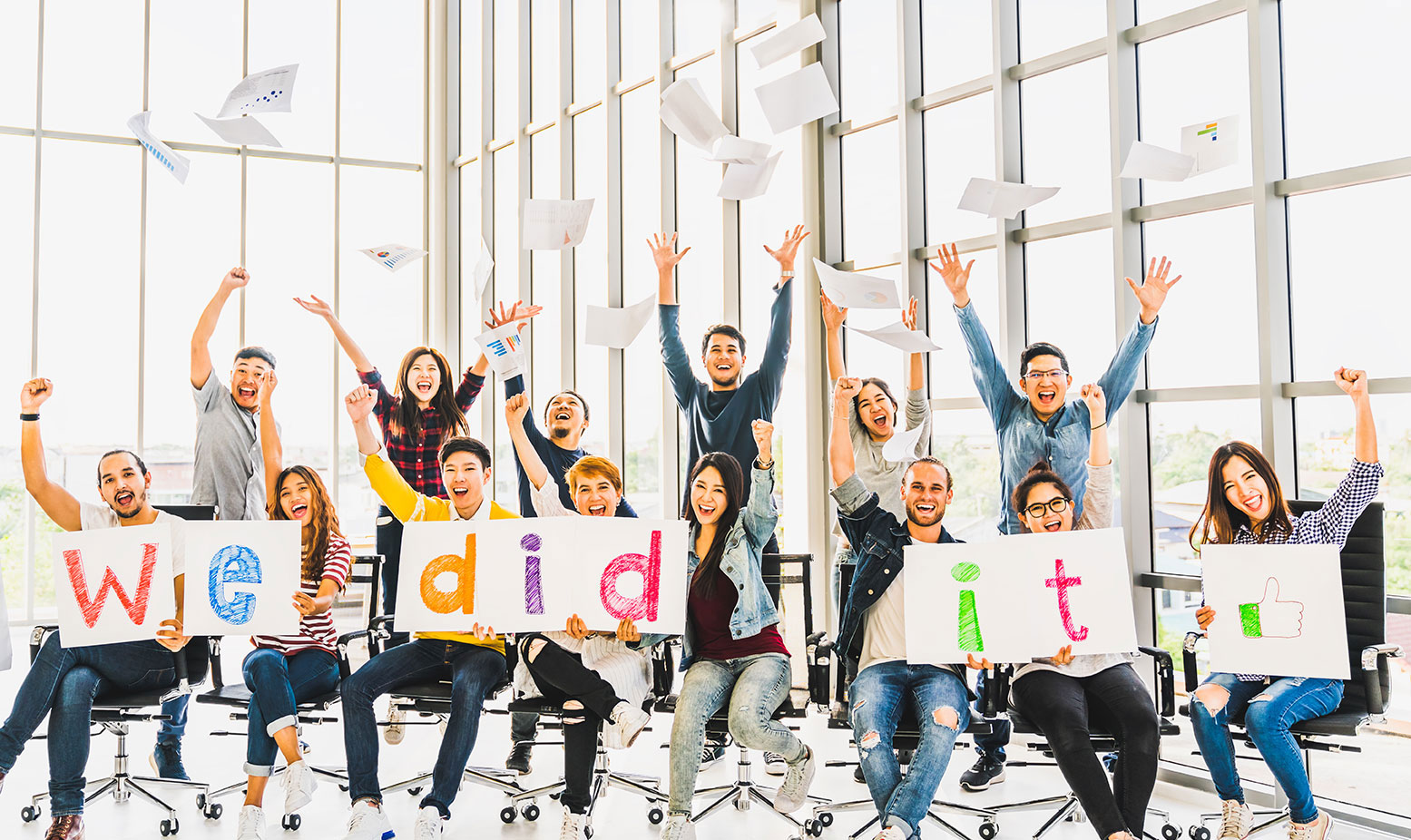 A group of people celebrate in an office, tossing papers and holding signs that spell “We did it!” with smiles and raised arms, excitedly marking their successful charter school renewal.